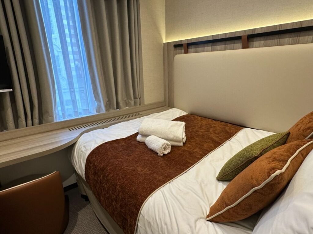 A neatly made hotel bed with brown and white linens, two pillows, and folded white towels on top showcases the fresh ambiance following Crown Group’s Mabledon Court Hotel Refurbishment Complete. There is a desk, chair, and a window with gray curtains letting in daylight.