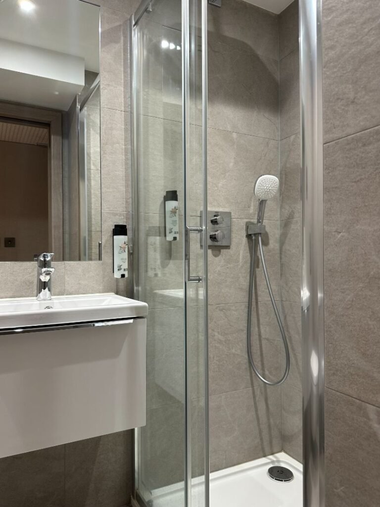 Modern bathroom with a glass shower enclosure, handheld showerhead, wall-mounted soap dispensers, grey tiled walls, and a white sink with a chrome faucet—a sleek example of the style featured in Crown Group’s Mabledon Court Hotel Refurbishment Complete.