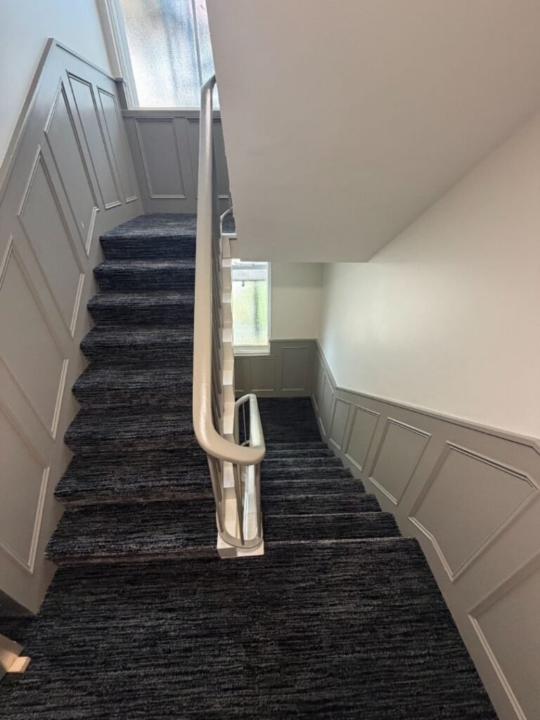 Carpeted staircase with gray paneling on the walls, a window at the landing, and a metal handrail running up the center, viewed from the top looking down—a refined detail of Crown Group’s Mabledon Court Hotel Refurbishment Complete.