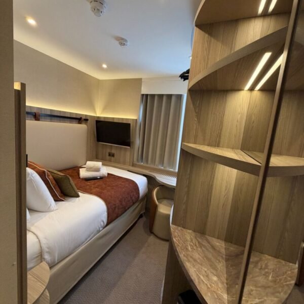 A modern hotel room at Mabledon Court Hotel features a single bed with a brown throw and plush pillows, a wall-mounted TV, and a window adorned with curtains. A unique wooden and marble shelving unit elegantly separates the sleeping area, while bright ceiling lights illuminate the space. Perfect for those seeking stylish comfort in hotels near Kings Cross or hotels in WC1H.