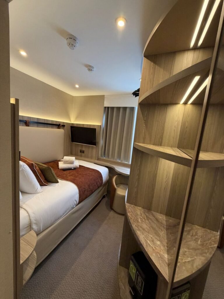 A modern hotel room at Mabledon Court Hotel features a single bed with a brown throw and plush pillows, a wall-mounted TV, and a window adorned with curtains. A unique wooden and marble shelving unit elegantly separates the sleeping area, while bright ceiling lights illuminate the space. Perfect for those seeking stylish comfort in hotels near Kings Cross or hotels in WC1H.