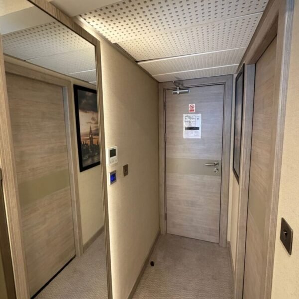 A hotel hallway in one of the stylish hotels near Kings Cross, with beige walls, a light wood door, a mirror on the left, a thermostat and blue button on the wall, and a notice posted on the door. The carpeted floor and perforated ceiling panels add to the modern feel commonly found in hotels in WC1H.