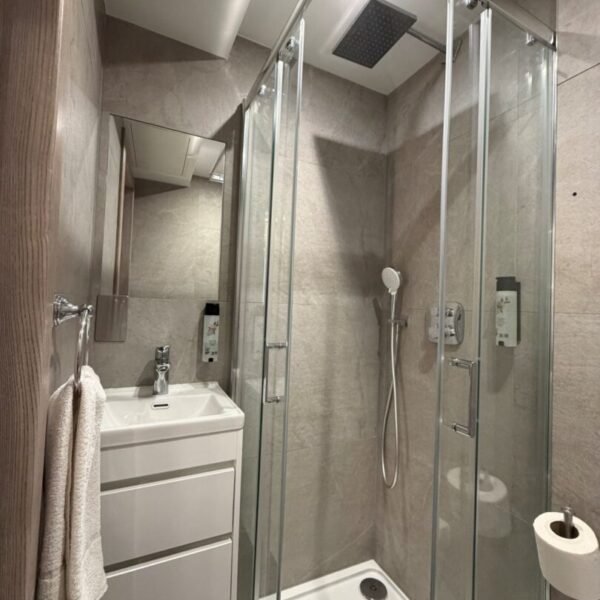 A modern, compact bathroom—like those found in top hotels near Kings Cross—features a glass shower enclosure, rainfall showerhead, handheld shower, white sink with cabinet, towel rack, and toilet paper holder on beige tiled walls.