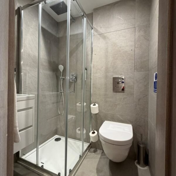 Modern bathroom with gray tiles, glass shower enclosure, wall-mounted toilet, two toilet paper rolls, and a small trash bin. Overhead lighting brightens the space, and fixtures are silver—reflecting the refined style you’d expect from top hotels in London like Mabledon Court Hotel.
