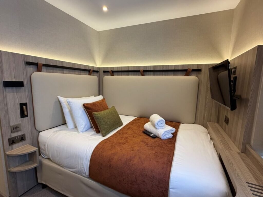 A small modern hotel room at Mabledon Court Hotel features a neatly made single bed with a brown throw and pillows, a towel set on the bed, and a wall-mounted TV facing the bed. The room offers soft lighting and minimalist decor, making it an ideal choice for travelers seeking hotels near Kings Cross in London.