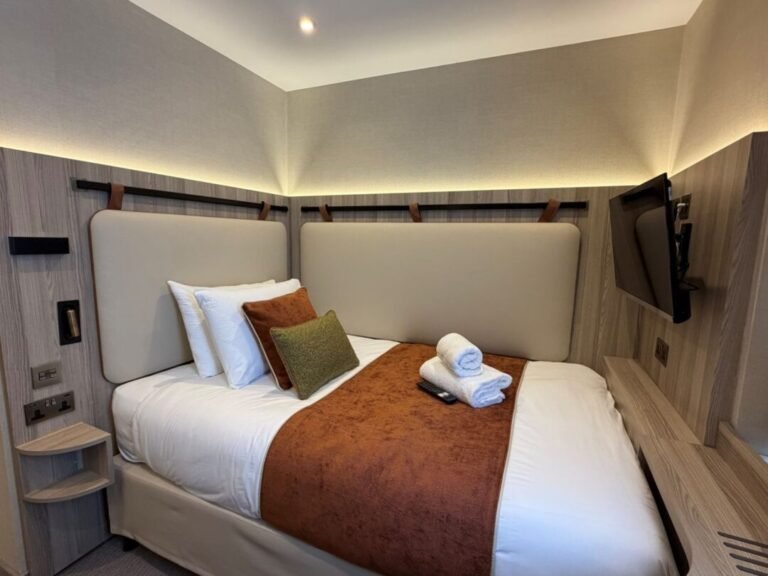 A small modern hotel room at Mabledon Court Hotel features a neatly made single bed with a brown throw and pillows, a towel set on the bed, and a wall-mounted TV facing the bed. The room offers soft lighting and minimalist decor, making it an ideal choice for travelers seeking hotels near Kings Cross in London.