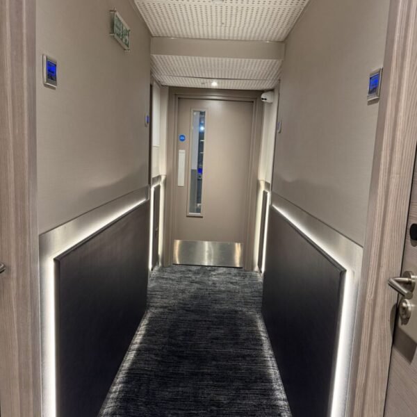 A modern, well-lit hallway with dark carpeting and wall panels, overhead lights, and an exit door at the end. An emergency exit sign is visible on the ceiling—typical of hotels in London such as Mabledon Court Hotel, conveniently located near Kings Cross.