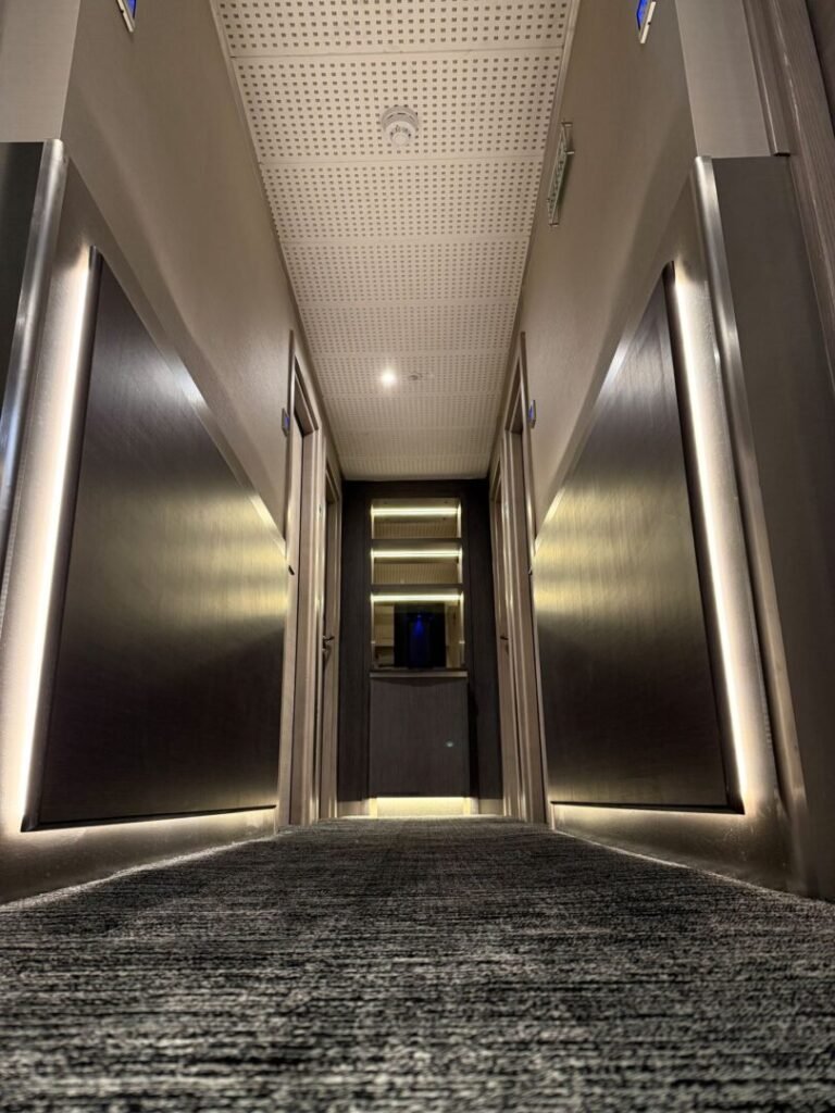 A low-angle view of a modern hotel hallway with carpeted floors, metallic wall panels, and soft ambient lighting along the walls, leading to a closed door at the end—an inviting scene often found in hotels near Kings Cross such as the renowned Mabledon Court Hotel and other stylish hotels in WC1H.