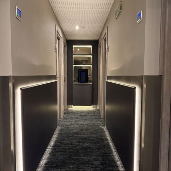 A modern, well-lit hallway at Mabledon Court Hotel features dark panels and light strips along the walls, a stylish grey carpet, and doors on both sides leading to a small alcove with a cabinet and mirror at the end—an inviting ambiance that sets this hotel apart among hotels near Kings Cross.