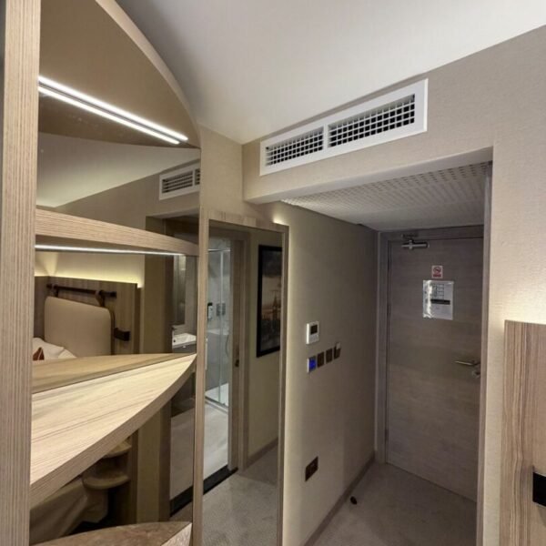 A modern hotel room corner at the Mabledon Court Hotel, one of the well-appointed hotels in WC1H, features a bunk bed on the left, a mirrored closet, beige walls, carpeted floor, and a closed door with ventilation and switches visible.