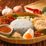 A woven basket filled with blue rice, sambal, fried chicken, crackers, half a boiled egg, peanuts, shredded coconut, and vegetables, surrounded by garlic, shallots, and red chilies on a bamboo mat.