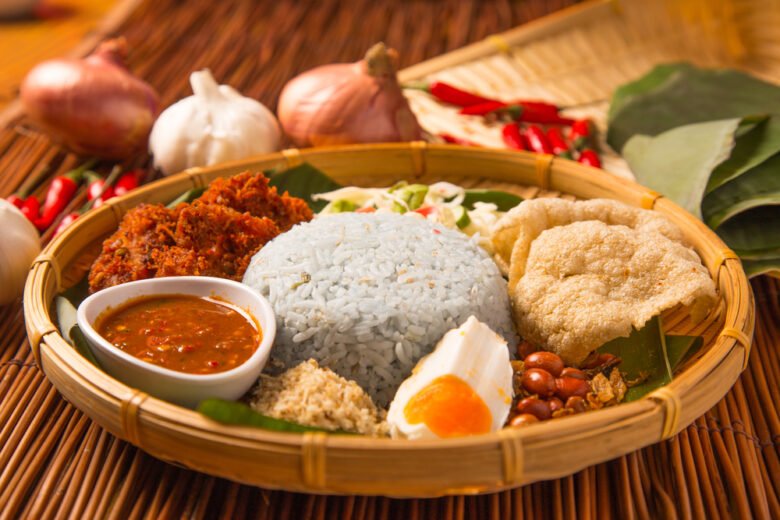 A woven basket filled with blue rice, sambal, fried chicken, crackers, half a boiled egg, peanuts, shredded coconut, and vegetables, surrounded by garlic, shallots, and red chilies on a bamboo mat.