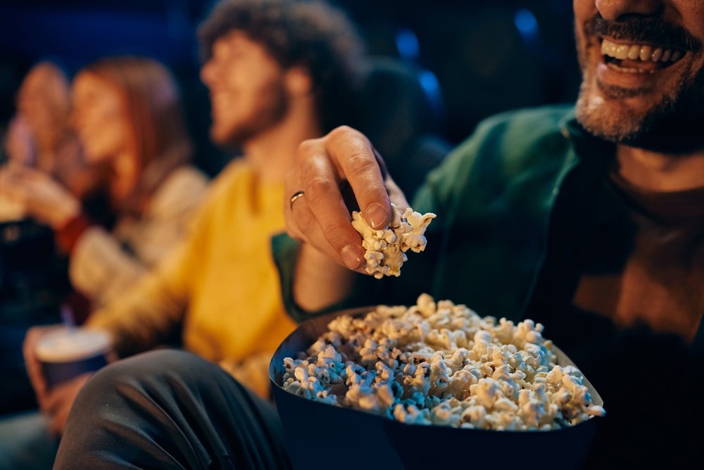 A group of people sit in a theater, smiling and enjoying popcorn. One person in the foreground is about to eat a handful of popcorn, while others are blurred in the background.