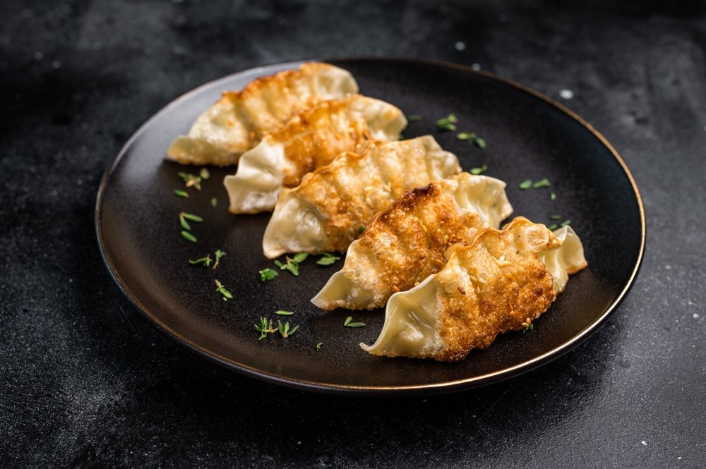 Four golden-brown pan-fried dumplings are arranged in a row on a dark plate, garnished with small green herb flakes, against a dark background.