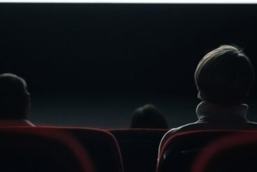 Three people sit in a dark movie theater, viewed from behind with the screen glowing softly in front of them. The focus is on the back of a person in the foreground with short hair and a light-colored collar.
