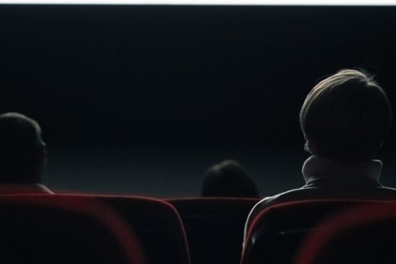 Three people sit in a dark movie theater, viewed from behind with the screen glowing softly in front of them. The focus is on the back of a person in the foreground with short hair and a light-colored collar.