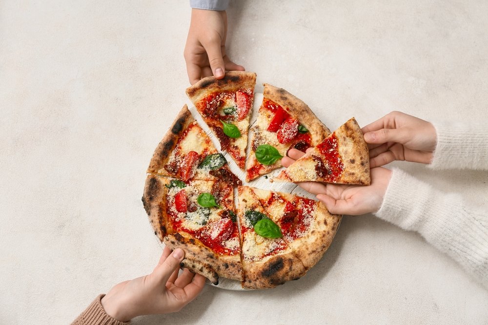 Three people reach for slices of a round pizza topped with tomato sauce, cheese, and basil leaves on a light background.