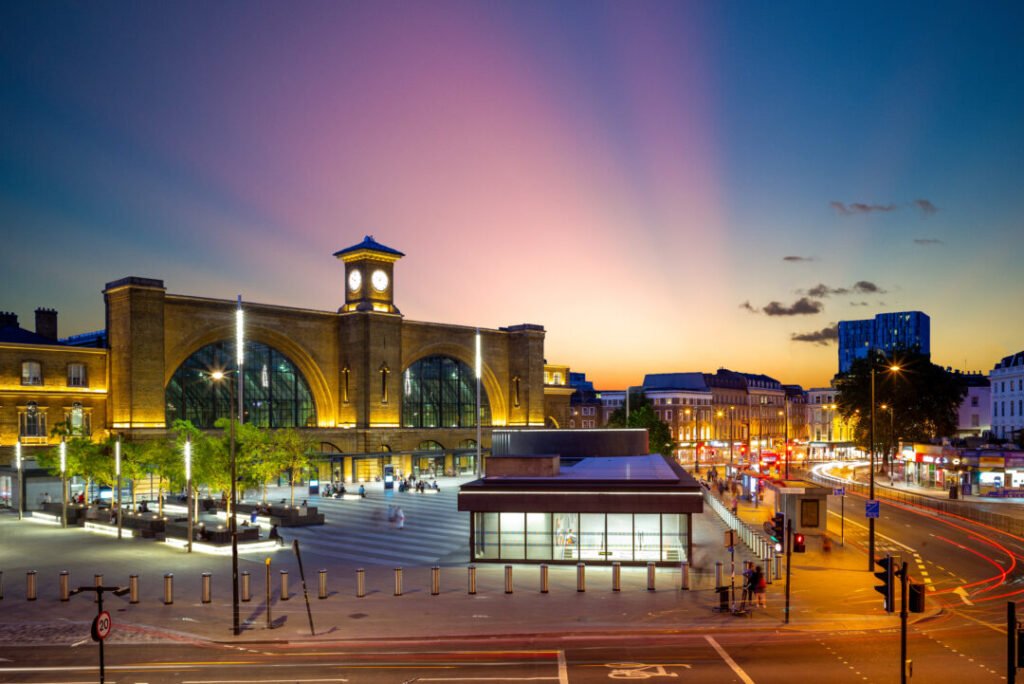 A view of London’s King’s Cross train station at sunset, with its illuminated clock tower, arched windows, nearby modern buildings, and light trails from traffic on busy streets.