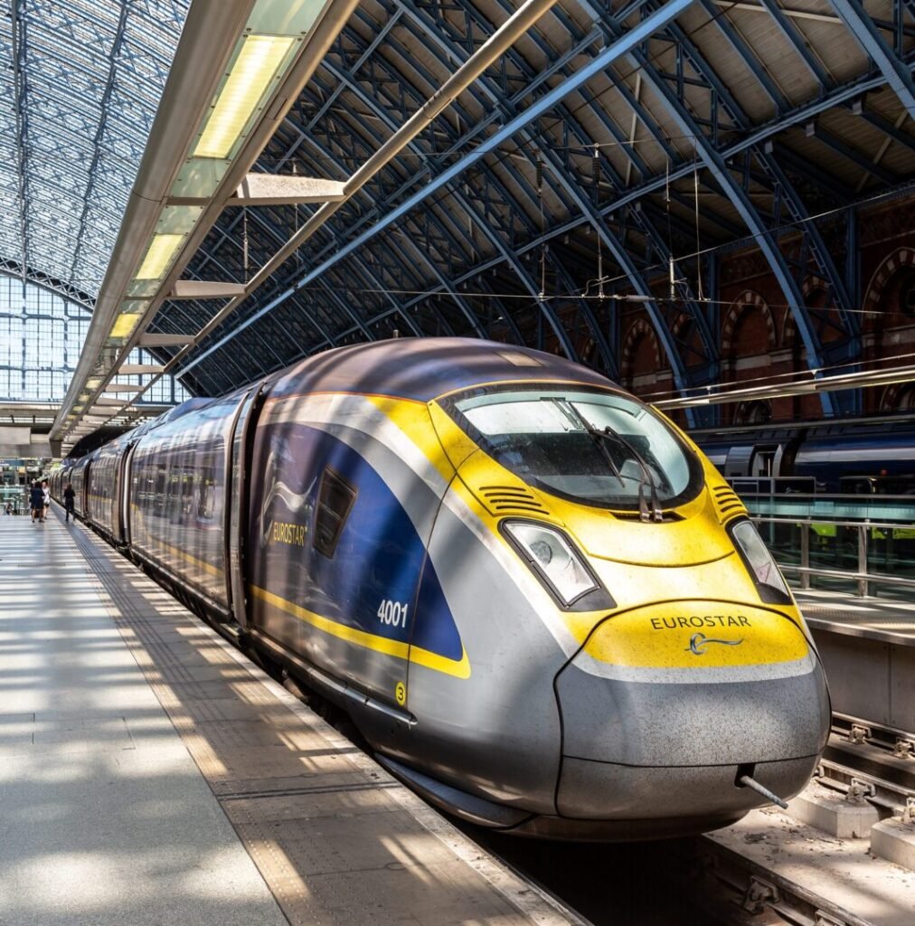 A Eurostar high-speed train with a yellow and blue design is stopped at a platform inside a large, modern glass-roofed train station.