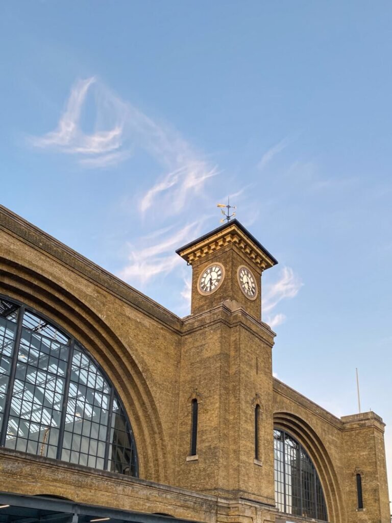 A brick clock tower with arched windows stands against a clear blue sky with wispy clouds. The clock shows nearly 6:45, and the building has a classic architectural style.