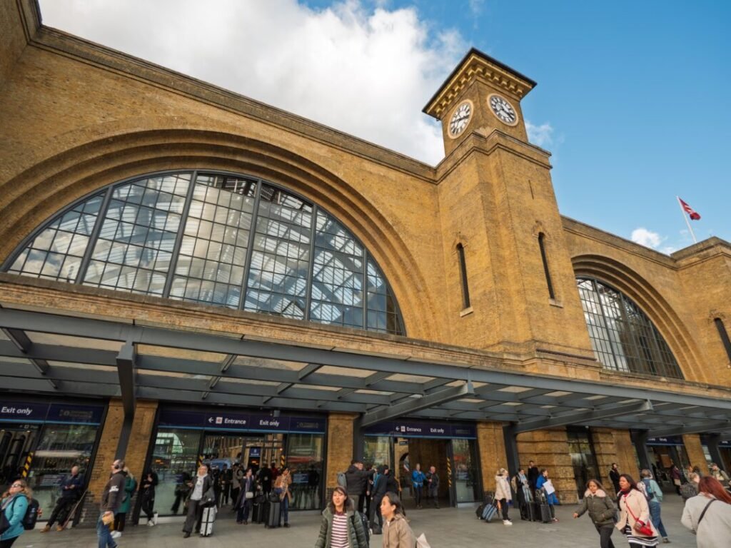 People stand and walk outside a large train station with arched windows and a clock tower under a blue sky with clouds. The building is made of brown brick, and there is a glass canopy above the entrances.