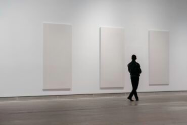 A person stands in an art gallery, observing three large, blank white canvases displayed on a plain white wall.