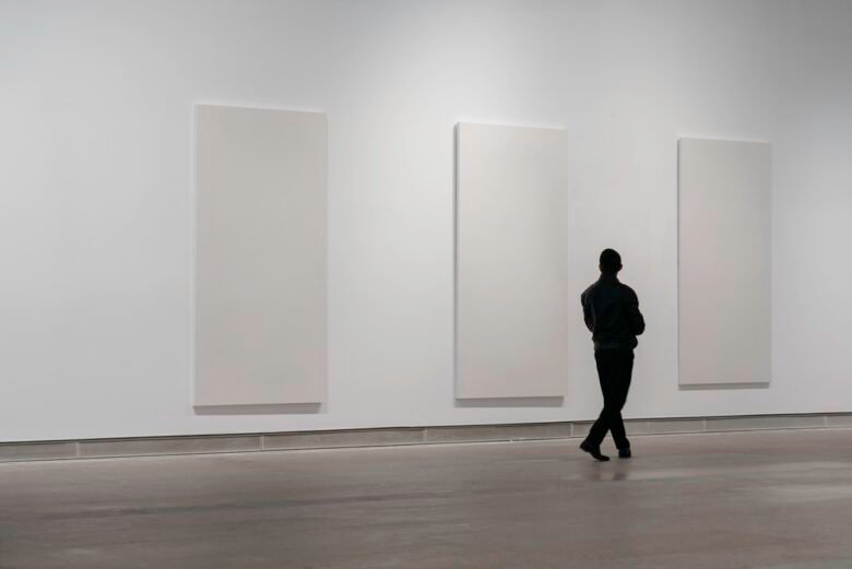 A person stands in an art gallery, observing three large, blank white canvases displayed on a plain white wall.
