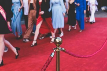 People in formal and semi-formal attire walk on a red carpet, with a gold stanchion and red rope in the foreground. The image focuses on legs and feet.