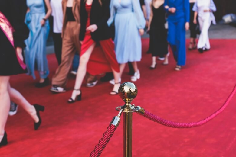 People in formal and semi-formal attire walk on a red carpet, with a gold stanchion and red rope in the foreground. The image focuses on legs and feet.