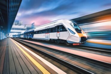A modern white and blue high-speed train moves quickly through a station, with motion blur visible in the background, suggesting rapid travel.