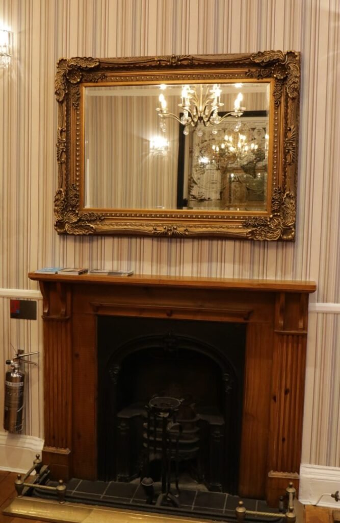 A wooden fireplace mantel with a large, ornate gold-framed mirror hanging above it against striped wallpaper.