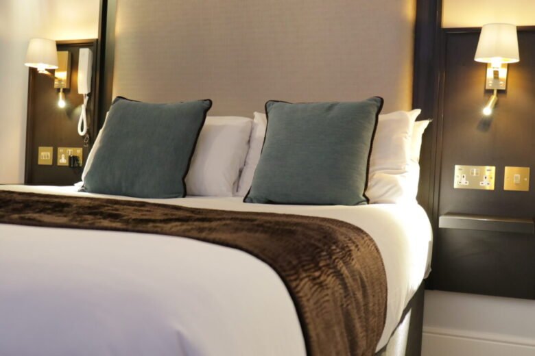 A made hotel bed with white linens, a dark brown runner, and two teal accent pillows, flanked by two wall-mounted lamps.