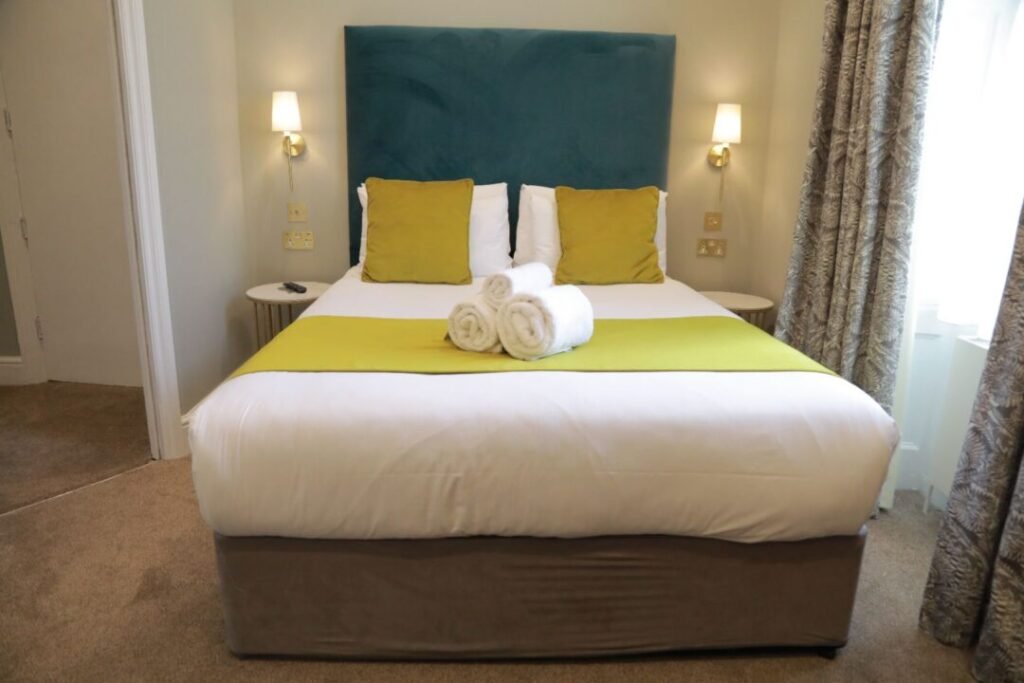 A hotel bed with a teal headboard, white linens, yellow accent pillows, and rolled white towels on the bed runner.