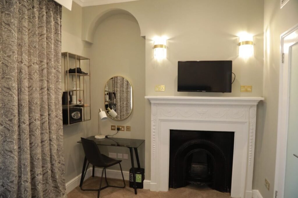 A hotel room featuring a desk with a chair and mirror next to a fireplace with a wall-mounted television above it.