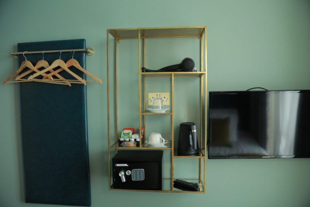 A metal wall shelf holding a hairdryer, tea and coffee supplies, a kettle, and a safe, positioned next to a wall-mounted television and a coat rack on a green wall.